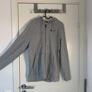 Grå hoodie från Nike - Säljer en grå hoodie från Nike med dragkedja och huva. Den har en klassisk design med Nike-logga på bröstet och är tillverkad i Dri-FIT-material för extra komfort. Perfekt för en avslappnad stil.
