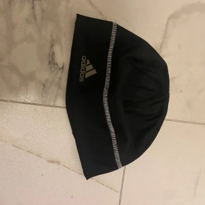 Svart mössa från Adidas - Snygg svart mössa från Adidas med en reflekterande rand och logotyp. Perfekt för kyliga dagar och sportaktiviteter. Tillverkad i ett mjukt och bekvämt material. Helt ny och använd