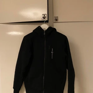 Svart zip från Sail Racing - Snygg svart zip från Sail Racing med dragkedja framtill. Perfekt för kyligare dagar med sin stilrena design och praktiska fickor. 