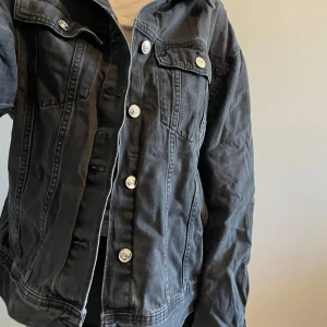 Svart jeansjacka från H&M - Snygg svart jeansjacka från H&M med knappar framtill och två bröstfickor med lock. Perfekt för en casual look och passar till det mesta. Klassisk design med krage och långa ärmar.