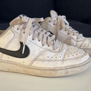 Vita Nike sneakers med svart swoosh - Säljer ett par vita Nike sneakers med svart swoosh. Skorna har snörning och perforerade detaljer på ovansidan för extra ventilation. Perfekta för en sportig look.