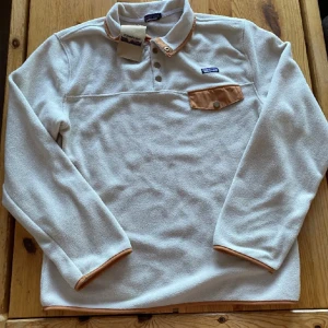 Vit fleece tröja från Patagonia - Selling a super cozy white vintage fleece sweater from Patagonia. It has a nice brown detail on the chest pocket and at the cuffs. Perfect for chilly days and has a classic collar with buttons. The shirt is in mint condition and still has the label. Perfect for fall and winter! new with tags