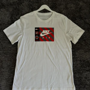 Vit Nike T-shirt med röd och svart logga - Säljer en snygg vit T-shirt från Nike med en cool röd och svart logga på framsidan. Perfekt för vardagsbruk eller träning. T-shirten är i bomull och har en normal passform. Kortärmad och superbekväm!