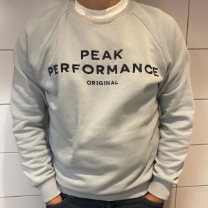 Grå sweatshirt från Peak Performance - Säljer en stilren och bekväm ljusblå sweatshirt från Peak Performance. Den har en klassisk design med svart text på framsidan och är perfekt för både vardag och träning. Tröjan har långa ärmar och ribbade muddar vid ärmslut och nederkant.