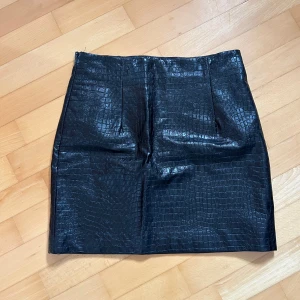 Svart kjol med krokodilmönster - Säljer en supercool svart kjol med krokodilmönster. Den är perfekt för en utekväll eller fest! Kjolen har en tight passform och är gjord av ett glansigt material som ger en lyxig känsla. Den har en dragkedja bak till för enkel på- och avtagning. Passar perfekt till en snygg topp och klackar!