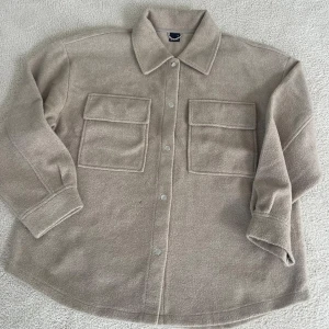 Beige overshirt från Ginatricot - Säljer en snygg beige overshirt från Ginatricot i storlek XL. Den är gjord i ett mjukt och mysigt material, perfekt för höst och vår. Skjortan har långa ärmar och två stora fickor framtill. Stängs med knappar och har en klassisk krage. Perfekt för lager-på-lager-stilen!