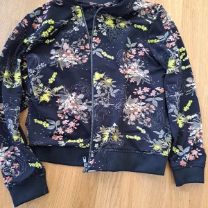 Blommig jacka med dragkedja - Säljer en superfin blommig jacka med dragkedja. Den har ett mörkt tyg med färgglada blommor i rosa, gul och grön. Perfekt för att liva upp garderoben! Jackan har långa ärmar och ribbade muddar vid ärmslut och nederkant. Passar perfekt för vår och höst.