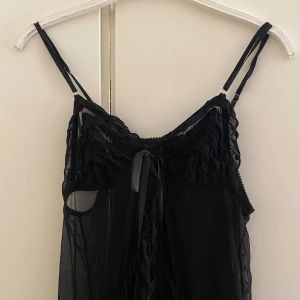 Svart transparent klänning med volanger - Säljer en superfin svart klänning med tunna axelband och volangdetaljer. Den är transparent och har en söt knytning framtill. Perfekt för en festlig kväll eller som ett lager över en annan outfit. Klänningen  ger en elegant och lite mystisk look. Den är i bra skick förutom ena bandet, syns på sista bilden!