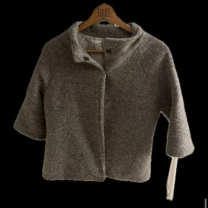 Grå ullkappa med trekvartsärm - Säljer en stilren grå ullkappa med trekvartsärm. Perfekt för höst och vår, med en elegant krage och knappar framtill. Kappan är i bra skick och ger en klassisk look. Passar både till vardags och festliga tillfällen.