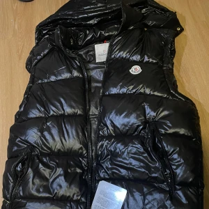 Svart dunväst från Moncler - PASSAR SOM M-L!! Säljer en snygg svart dunväst från Moncler i nytt skick. Västen har en glansig yta och är quiltad för extra värme. Den har en dragkedja framtill och praktiska fickor. Perfekt för höst och vinter! 🖤