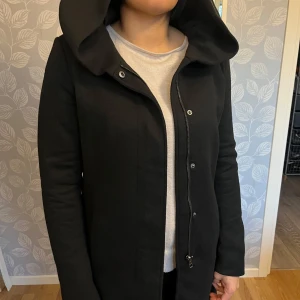 Svart kappa med huva - Säljer en stilren svart kappa med huva. Den har en dragkedja och knappar framtill för extra skydd mot kyla. Perfekt för höst och vinter, med en elegant och tidlös design.