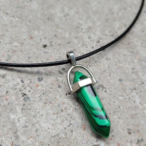 Halsband med grön sten i glas - Snyggt halsband med en grön och svart mönstrad glassten. Hänget är format som en spetsig kristall och hänger på en tunn svart rem. Perfekt för att ge en färgklick till din outfit! Hänget är i glas. Remmens längd ca 45cm.