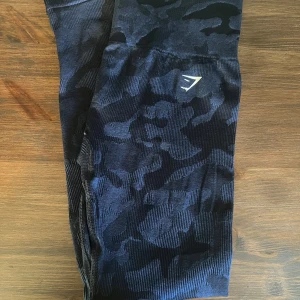 Blå camo leggings från Gymshark - Säljer ett par svarta camo leggings från Gymshark i storlek S. De är perfekta för träning med sin tighta passform och bekväma material. Leggingsen har en snygg camouflagemönster och är i mycket bra skick. Perfekta för gymmet eller en aktiv vardag!