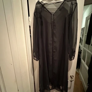 Svart transparent neglige(?) - Säljer en elegant svart neglige i transparent material. Klänningen har långa ärmar och en v-ringad design med en liten dekorativ knapp om man vill knäppa. 