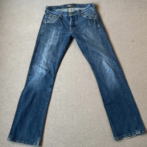 Lågmidjade Blå jeans från Lee bootcut - Säljer ett par klassiska blå jeans från Lee .  tillverkade i bomull, vilket gör dem både bekväma och hållbara. Perfekta för vardagsbruk och kan enkelt matchas med olika outfits. Jeansens skick är mycket bra och de har en traditionell knappgylf.
