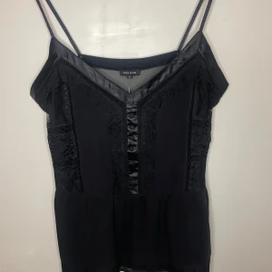 Svart topp från River Island - Snygg svart topp från River Island i storlek 38. Den har smala axelband och fina spetsdetaljer framtill. Perfekt för en utekväll eller fest! Toppen är i mycket bra skick och ger en elegant look.