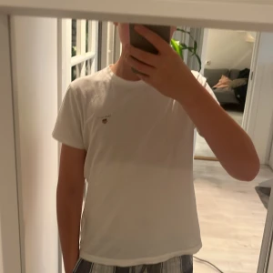Vit t-shirt från GANT - Säljer en klassisk vit t-shirt från GANT i mycket bra skick. Den har en liten logga på bröstet som ger en stilren look. Perfekt för vardagsbruk eller sommarens äventyr. Passar till allt och är superbekväm!