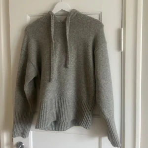 Grå stickad hoodie - Mysig grå stickad hoodie perfekt för kyliga dagar. Den har en avslappnad passform med långa ärmar och en ribbad nederkant som ger extra stil. Huvan har snören för justering. Perfekt för både höst och vinter!