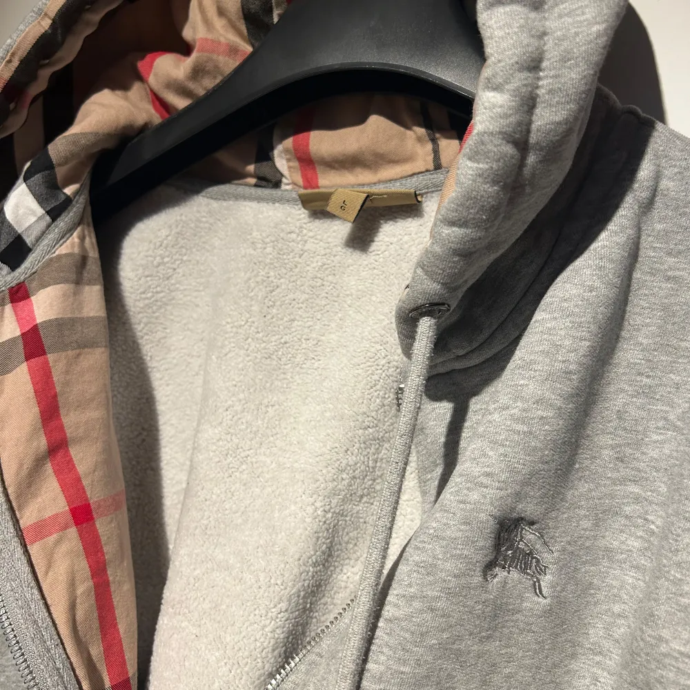 Säljer en grå hoodie från burberry med dragkedja och ett snyggt rutigt foder i huvan. Perfekt för höst och vår, med långa ärmar och en broderad logga på bröstet. Den är i bra skick och superbekväm. Passar perfekt för en casual look eller när du bara vill mysa hemma. L men passar M. Hör av er vid minsta fundering 🍾. Tröjor & Koftor.