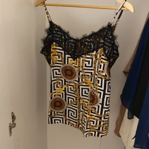 Mönstrad linne med spetsdetaljer - Säljer en snygg linne med ett unikt mönster i svart, vitt och guld. Den har tunna axelband och vacker spets längs halsringningen. Perfekt för sommardagar eller en utekväll. Passar bra till både jeans och kjol!