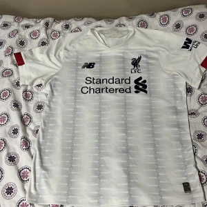 Vit Liverpool fotbollströja  - Säljer en vit Liverpool fotbollströja från New Balance i mycket bra skick. Den har Liverpool FC-loggan och Standard Chartered som sponsortryck. Perfekt för alla Liverpool-fans! 🔥ingen aning om den är äkta eller inte 
