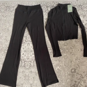 Svart träningsset med byxor och jacka - Snyggt svart träningsset med byxor och  matchande jacka. Byxorna har en bootcut-stil och är perfekta för både träning och vardag. Jackan har en dragkedja framtill och långa ärmar. Setet är gjort i ett mjukt och stretchigt material för maximal komfort. Som ny prislapp på