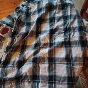 Rutig skjorta från KappAhl stl L (43/44) - Säljer en snygg rutig skjorta från KappAhl i svart, vit och grå. Skjortan har korta ärmar och knappar framtill. Perfekt för både vardag och fest! Den är i bra skick och passar till många olika stilar.