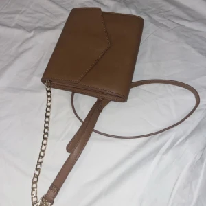 Brun axelväska med kedja - Snygg brun axelväska i syntetmaterial med en elegant kedjedetalj på axelremmen. Väskan har ett kuvertliknande lock och flera fack inuti för bra organisering. Perfekt för både vardag och fest!