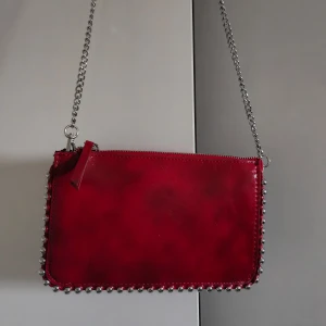 Röd axelväska med kedja - Snygg röd axelväska i skinn med silverfärgad kedja och coola nitar längs kanterna. Perfekt för en utekväll eller fest! Väskan har en dragkedja upptill för säker förvaring av dina saker.