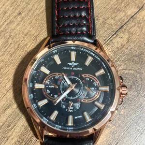 Svart och roséguld klocka från Geneva Design - Snygg klocka från Geneva Design med svart urtavla och detaljer i roséguld. Den har en tachymeter och flera små urtavlor för extra funktioner. Armbandet är i svart skinn med röda och vita sömmar. Perfekt för både vardag och fest!
