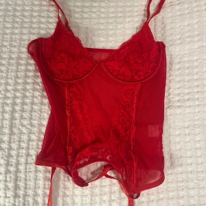 Röd spetskorsett - Snygg röd korsett med spetsdetaljer och justerbara axelband. Perfekt för en festlig kväll eller speciella tillfällen. Den har en tight passform och är gjord i ett syntetmaterial som ger en smickrande siluett. Fästena nedtill gör den extra elegant. 