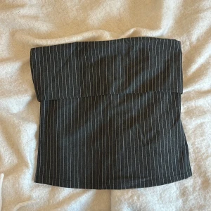 Randig tubtopp från Stories - Snygg och stilren tubtopp från Stories i storlek M. Den är mörkgrå med tunna vita ränder och har en dragkedja bak för enkel påklädning. Perfekt för en chic och modern look!