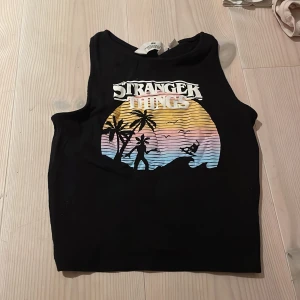 Svart Stranger Things linne från H&M - Coolt svart linne från H&M med Stranger Things-tryck i retrostil. Perfekt för fans av serien! Linnet är ärmlöst och har en avslappnad passform. Materialet är mjukt och bekvämt, perfekt för sommardagar eller som en del av en lager-på-lager-look.