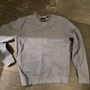 Grå sweatshirt från Lyle & Scott - Säljer en stilren lite gråblå sweatshirt från Lyle & Scott i mycket bra skick. Tröjan har en klassisk rund halsringning och det ikoniska gula logotypmärket på bröstet. Perfekt för både vardag och avslappnade tillfällen. Passar bra till jeans eller chinos!
