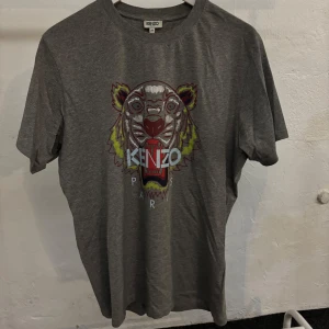 Grå t-shirt med tigertryck från Kenzo - Säljer en grå t-shirt från Kenzo i storlek M. Den har ett coolt tigertryck i flera färger på framsidan och är perfekt för vardagsbruk. T-shirten är i bra skick och gjord av mjukt material. Passar perfekt till jeans eller shorts!