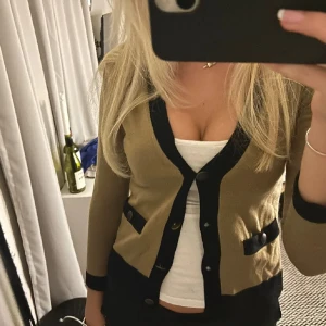 Filippa K kofta - Snygg beige kofta från Filippa K med svarta detaljer och knappar. Perfekt för både vardag och fest. Den har en klassisk look med långa ärmar och en skön passform. Passar bra över en topp eller klänning. Bild från förra säljaren.