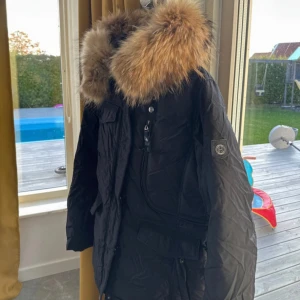 Svart vinterjacka med pälsdetaljer - Säljer en snygg svart parkajacka med stor päls på huvan. Perfekt för kalla vinterdagar. Jackan har flera fickor och en dragkedja framtill. Den är i mycket bra skick och redo för nya äventyr!
