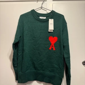 Grön stickad tröja från Ami - Säljer en snygg grön stickad tröja från Ami med ett rött hjärta och bokstaven A på bröstet. Tröjan har en klassisk rund hals och långa ärmar. Perfekt för att hålla sig varm och stilren under kyliga dagar. Helt ny med taggar kvar!
