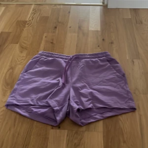 Lila shorts från Amisu - Säljer ett par lila shorts från Amisu i mycket bra skick. De är tillverkade i ett mjukt material och har en elastisk midja med dragsko för justerbar passform. Perfekta för sommarens varma dagar eller en avslappnad dag hemma. Storlek EU 34.