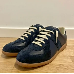 Blå sneakers från Maison Margiela - Snygga blå sneakers från Maison Margiela med vit snörning och brun sula. Skorna har en stilren design med en kombination av mocka och skinnmaterial. Perfekta för en trendig look.