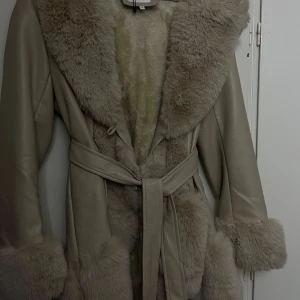 Beige kappa med pälsdetaljer - Snygg beige kappa från Cappuccini med mjuka pälsdetaljer på krage, ärmslut och insida. Jackan har ett bälte i midjan för en figursydd look och långa ärmar. Perfekt för kyligare dagar när du vill hålla stilen.