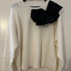 Vit tröja med svart rosett från Kappahl - stilren vit tröja från Kappahl med en stor svart rosett på axeln. Tröjan har en rund halsringning och långa ärmar, perfekt för en elegant look. Passar bra till både vardag och speciella tillfällen.