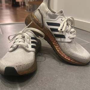 Adidas Ultraboost i vitt och brons - Snygga Adidas Ultraboost sneakers i vitt med bronsfärgad sula och svarta detaljer. Skorna har en stickad ovandel och snörning för optimal passform. Perfekta för både träning och vardag. Använda men funkar fortfarande lika bra som helt nya. 
