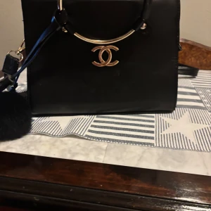 Svart handväska  - Elegant svart handväska med gulddetaljer och ett ikoniskt märke framtill. Väskan har en praktisk dragkedja och en fluffig dekoration. Perfekt för att bära dina nödvändigheter med stil. Knappt använt 