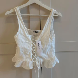 Vit linnetopp med snörning  - Säljer en söt vit croppad linnetopp från Gina Tricot med snörning framtill och volangdetalj vid nederkanten. Påminner om en korsettopp! Toppen är ärmlös och perfekt för varma dagar. Helt oanvänd!