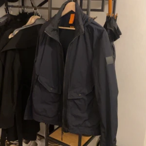 Mörkblå jacka från Boss - Säljer en stilren mörkblå jacka från Boss med dragkedja och huva. Jackan har praktiska fickor med knappar och en diskret logga på ärmen. Perfekt för kyligare dagar.
