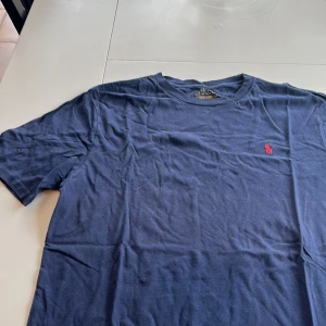 Mörkblå t-shirt från Ralph Lauren - Säljer en klassisk mörkblå t-shirt från Ralph Lauren med det ikoniska röda broderade logotypen på bröstet. T-shirten är kortärmad och tillverkad i mjuk bomull. Perfekt för en stilren och avslappnad look.