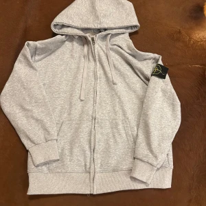 Grå hoodie från Stone Island - Säljer en stilren grå hoodie från Stone Island med dragkedja och justerbar huva. Tröjan har en ikonisk logga på ärmen och ribbade muddar vid ärmslut och nederkant. Perfekt för en avslappnad stil.