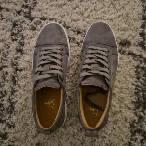 Grå sneakers med röd sula (christan louboutin) - Snygga grå sneakers med en kontrasterande röd sula och vita kanter. Skorna har snörning och är tillverkade i mocka, vilket ger en stilren och modern look. Perfekta för en avslappnad stil.