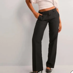 Svarta kostymbyxor - Snygga svarta kostymbyxor med hög midja och rak passform. Byxorna har en klassisk design med knappar och dragkedja framtill. Dom är helt nya och är kvar i plasten. Dom är från nelly!!